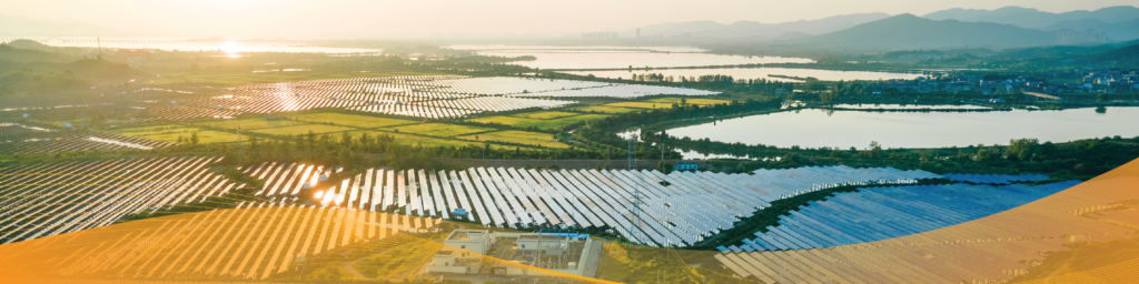 A wide panoramic banner showing a large utility-scale solar farm with rows of photovoltaic panels stretching across the landscape beside lakes and wetlands at sunset, with distant mountains on the horizon and a translucent orange graphic overlay along the bottom of the image.