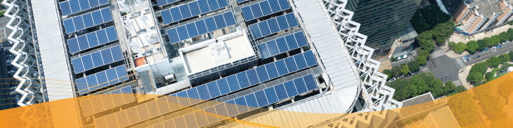 Aerial view of a commercial building with rooftop solar panels covering multiple sections of the roof, surrounded by neighboring buildings, streets, and trees, with a translucent orange graphic overlay along the bottom of the image.