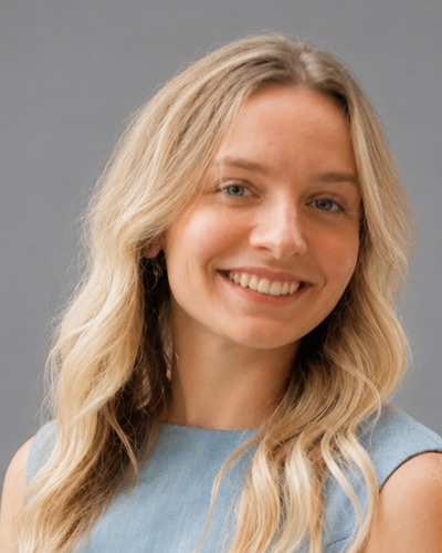 Headshot of a smiling person with shoulder-length wavy blonde hair, wearing a light blue sleeveless top, facing the camera against a soft gray studio background.