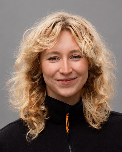 Headshot of a smiling person with shoulder-length curly blonde hair, wearing a black zip-up top, facing the camera against a soft gray studio background.